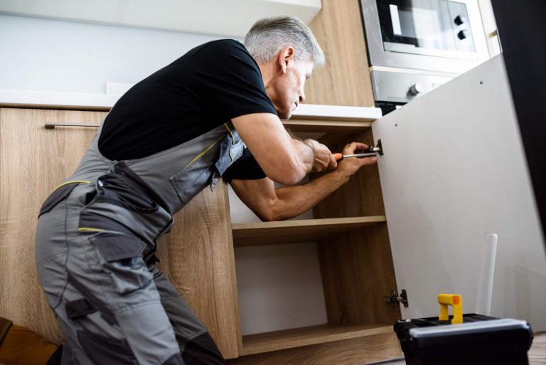 Aged repairman in uniform working, fixing kitchen cabinet using screwdriver. Repair service concept. Selective focus on person. Horizontal shot. Side view